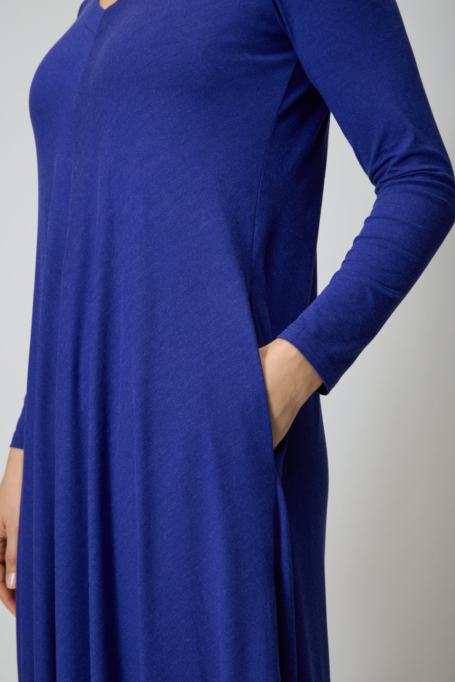 A person models the Soft Viscose Jersey Bubble Dress in blue, featuring long sleeves, a subtle bubble silhouette, and side pockets, standing slightly turned from the camera.