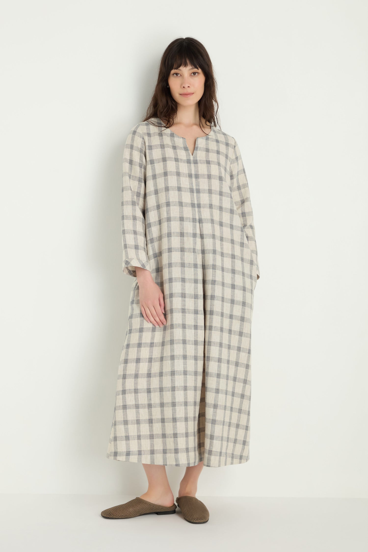 A woman stands against a white background wearing the Grid Check Linen Dress, a loose-fitting, long-sleeve linen dress with a grid check pattern, paired with brown slip-on shoes.