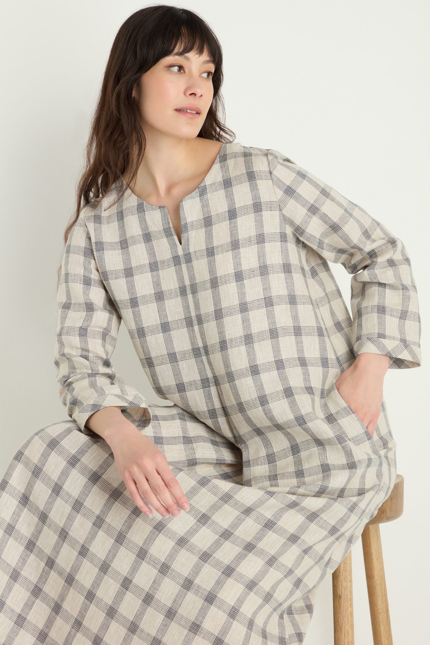 A woman sits on a wooden stool against a plain background, looking to the side, wearing the Grid Check Linen Dress—a long-sleeved linen dress in neutral tones with a grid pattern.