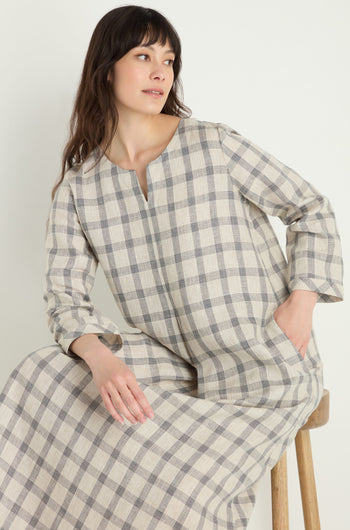 A woman sits on a wooden stool against a plain background, looking to the side, wearing the Grid Check Linen Dress—a long-sleeved linen dress in neutral tones with a grid pattern.