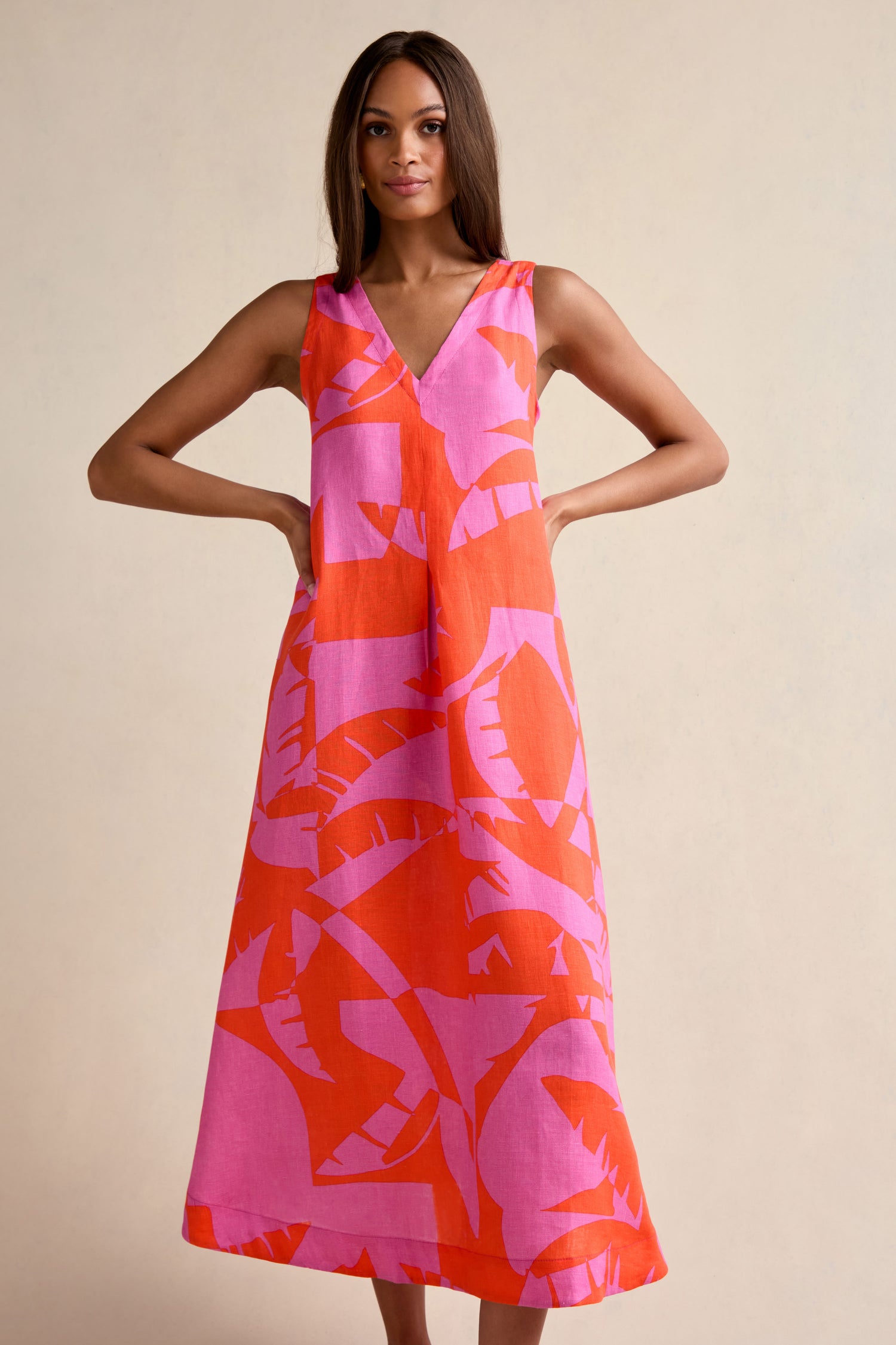 A woman models the Palm Mirage Linen Flared Dress, a sleeveless, ankle-length fit-and-flare style featuring a pink and orange abstract leaf pattern, set against a plain beige background.