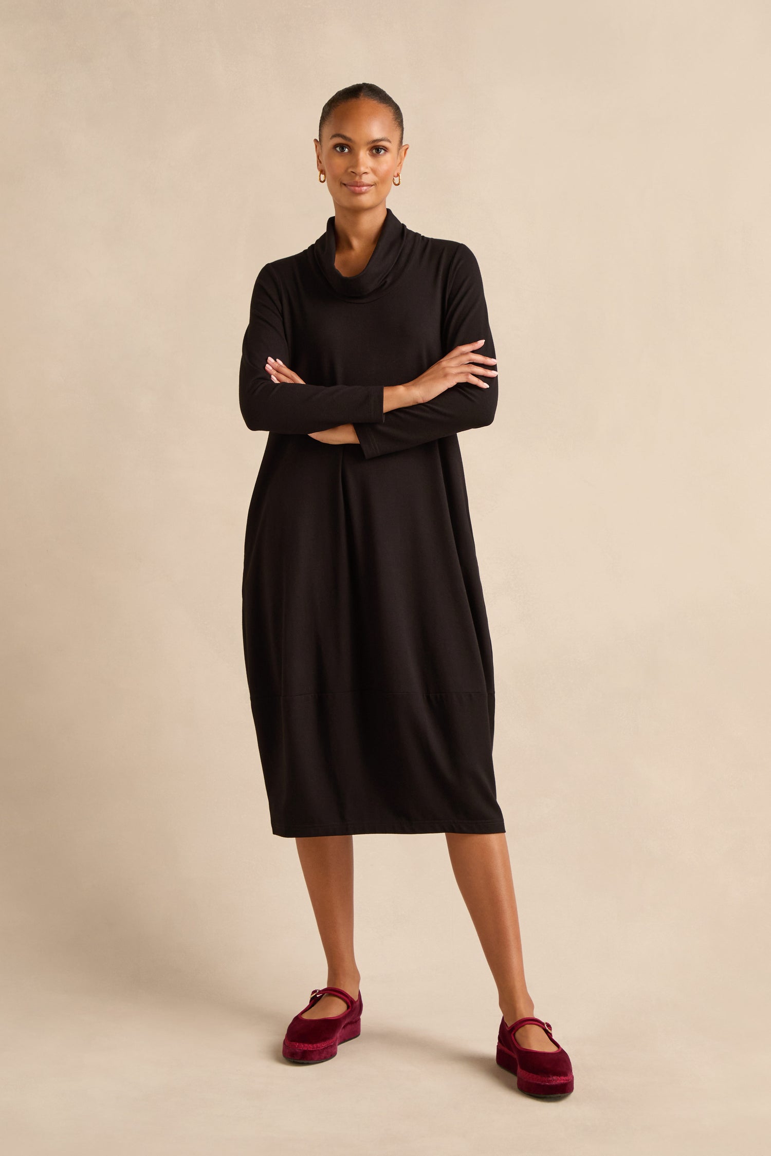Woman standing with arms crossed, wearing the Fluid Crepe Bubble Dress in black and red velvet shoes, against a neutral beige background.