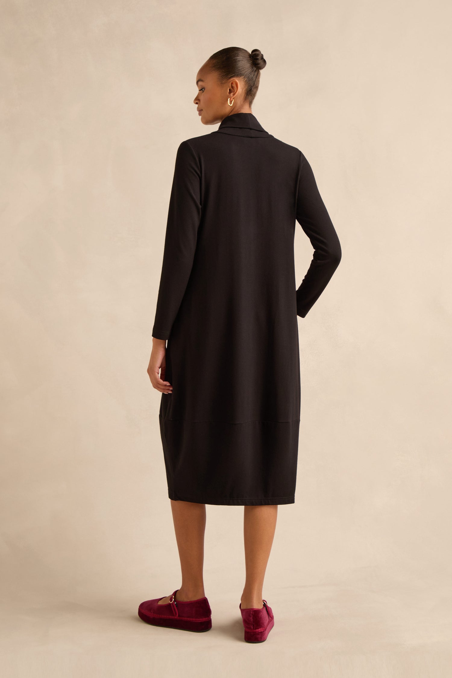 A woman stands against a neutral background, facing away with one hand on her hip, wearing the Fluid Crepe Bubble Dress—a long-sleeve, black knee-length design crafted from fluid crepe—paired with red velvet loafers.