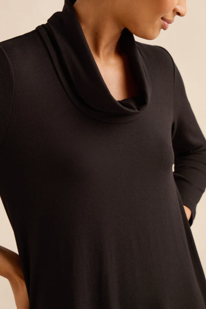 A person models the Fluid Crepe Bubble Dress in black, featuring long sleeves and a cowl neckline, shown from shoulder to waist with hands on hips against a neutral backdrop.
