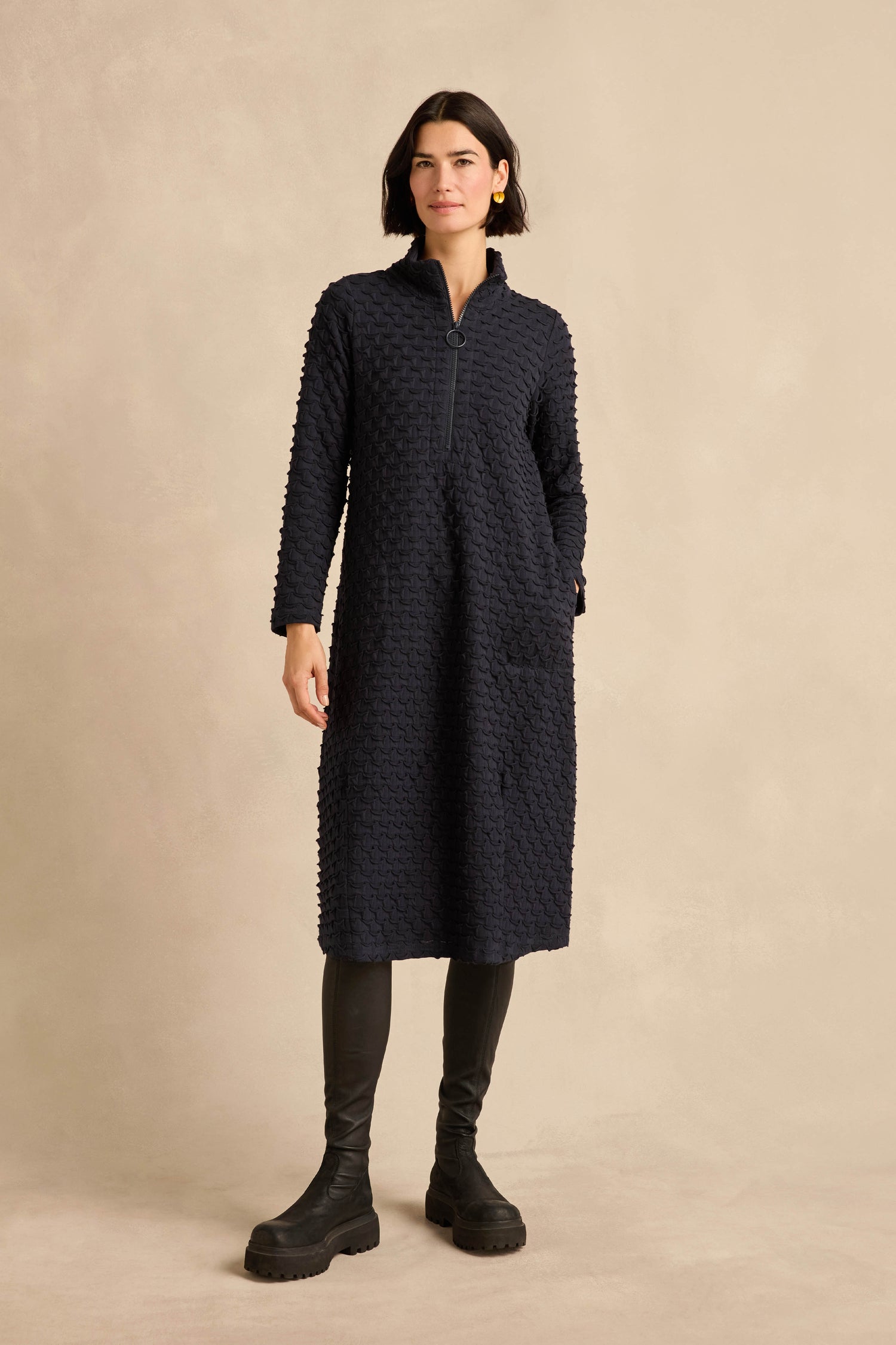 Woman wearing the Origami Pocket Jersey Zip Neck Bubble Dress in navy, featuring a textured jersey fabric, long sleeves, a zip-neck collar with pocket detail, paired with black leggings and chunky black boots, standing against a beige background.