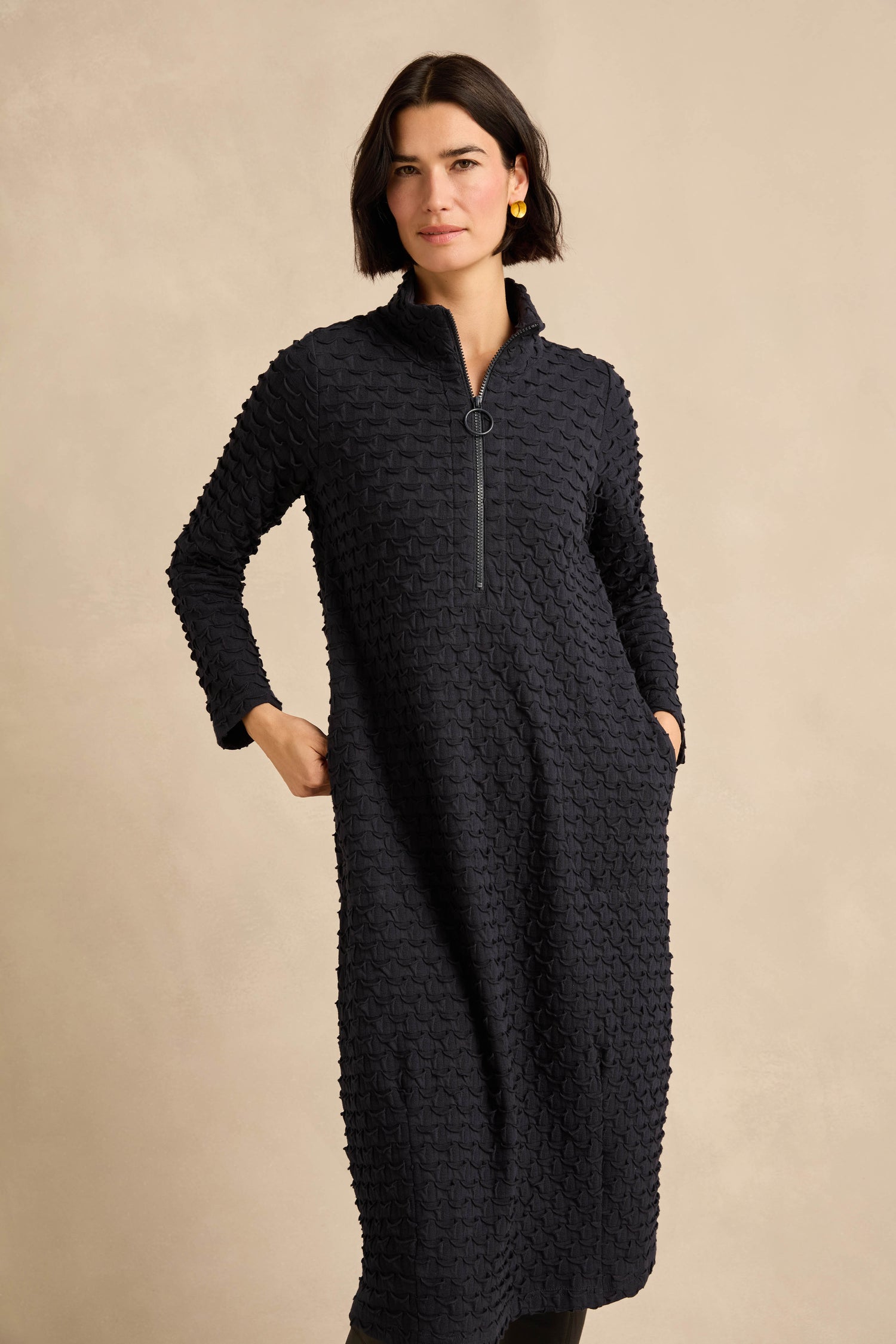 A woman stands with her hands in the pockets of the Origami Pocket Jersey Zip Neck Bubble Dress—featuring long sleeves, textured black jersey fabric, and a zip neck with a half-zip front—set against a beige background.