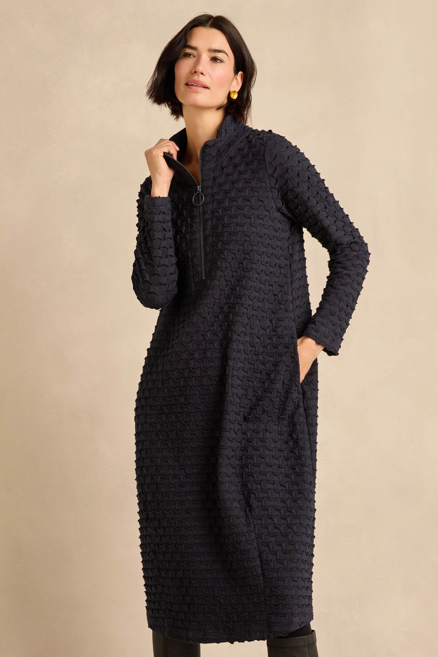 A woman stands against a neutral background wearing the Origami Pocket Jersey Zip Neck Bubble Dress—a long-sleeved, dark-colored midi dress with a textured jersey fabric, zip neck collar, and pockets. Her right hand is near her chin while her left hand rests in a pocket.