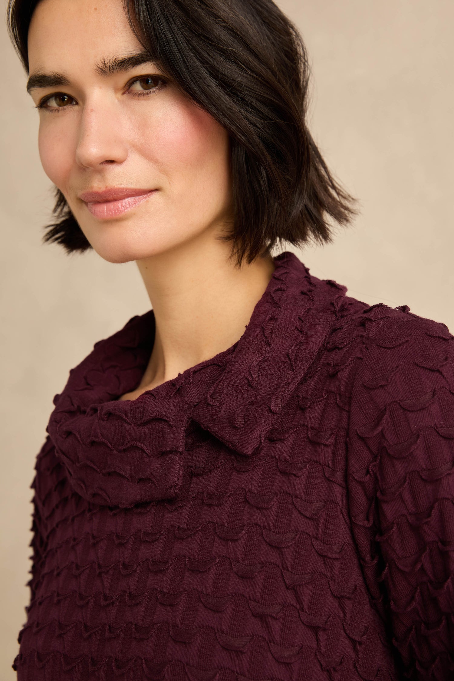 A woman with short dark hair wears the Origami Pocket Jersey Cowl Neck Dress in deep purple, featuring a textured fabric and wide collar, as she looks at the camera with a neutral expression.