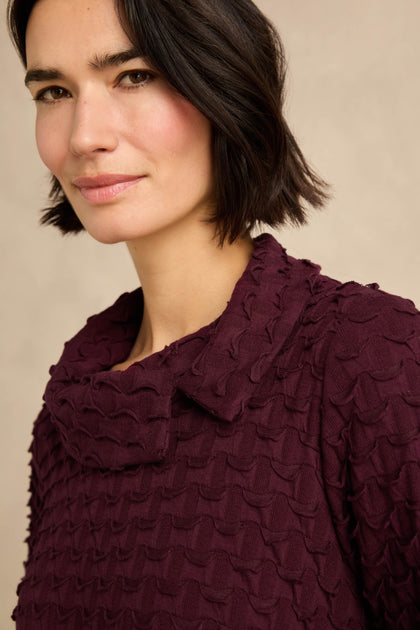 A woman with short dark hair wears the Origami Pocket Jersey Cowl Neck Dress in deep purple, featuring a textured fabric and wide collar, as she looks at the camera with a neutral expression.