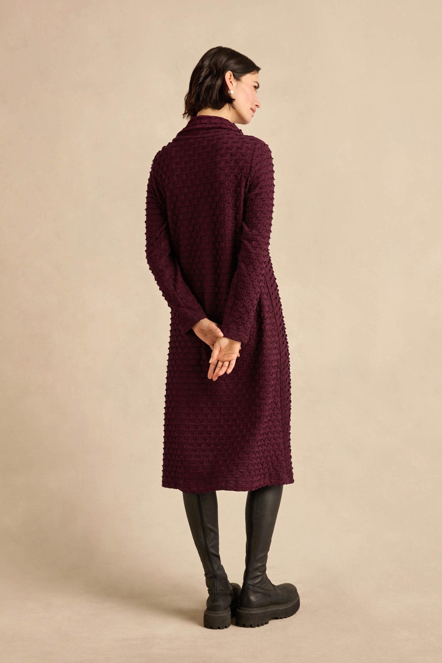 A person stands with their back to the camera, hands clasped behind them, wearing the Origami Pocket Jersey Cowl Neck Dress in textured maroon and black boots, set against a neutral beige background.