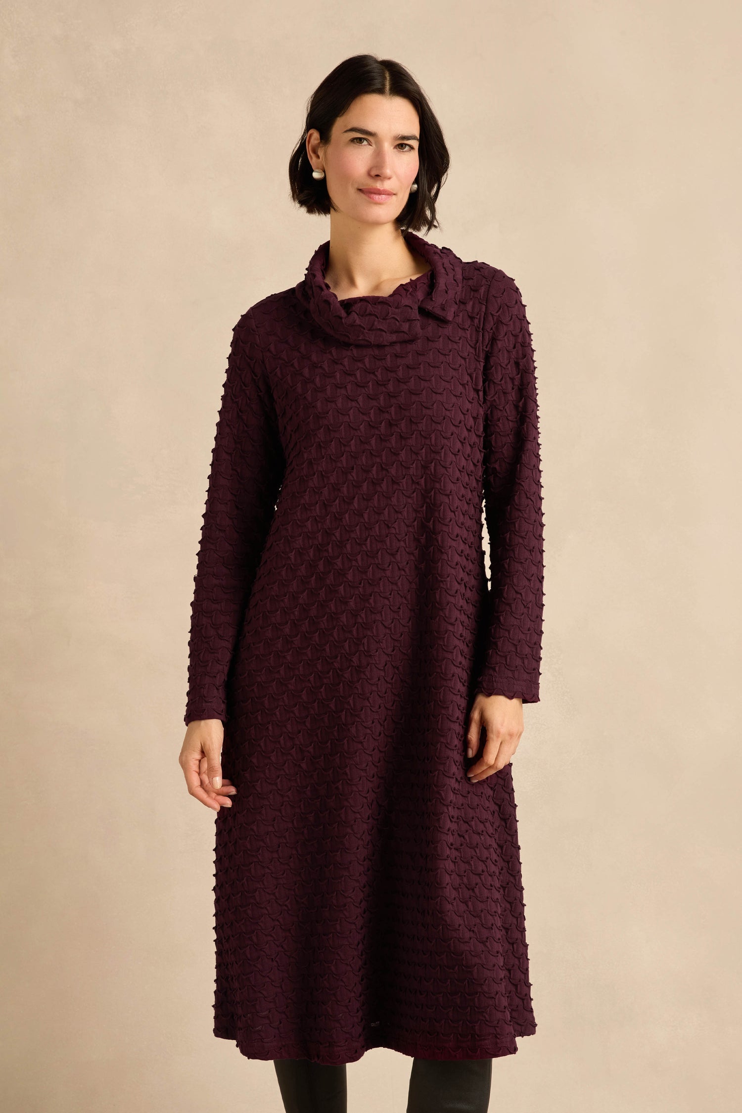 A woman wearing the Origami Pocket Jersey Cowl Neck Dress, featuring long sleeves and a textured jersey fabric in a deep plum hue, stands against a plain beige background.