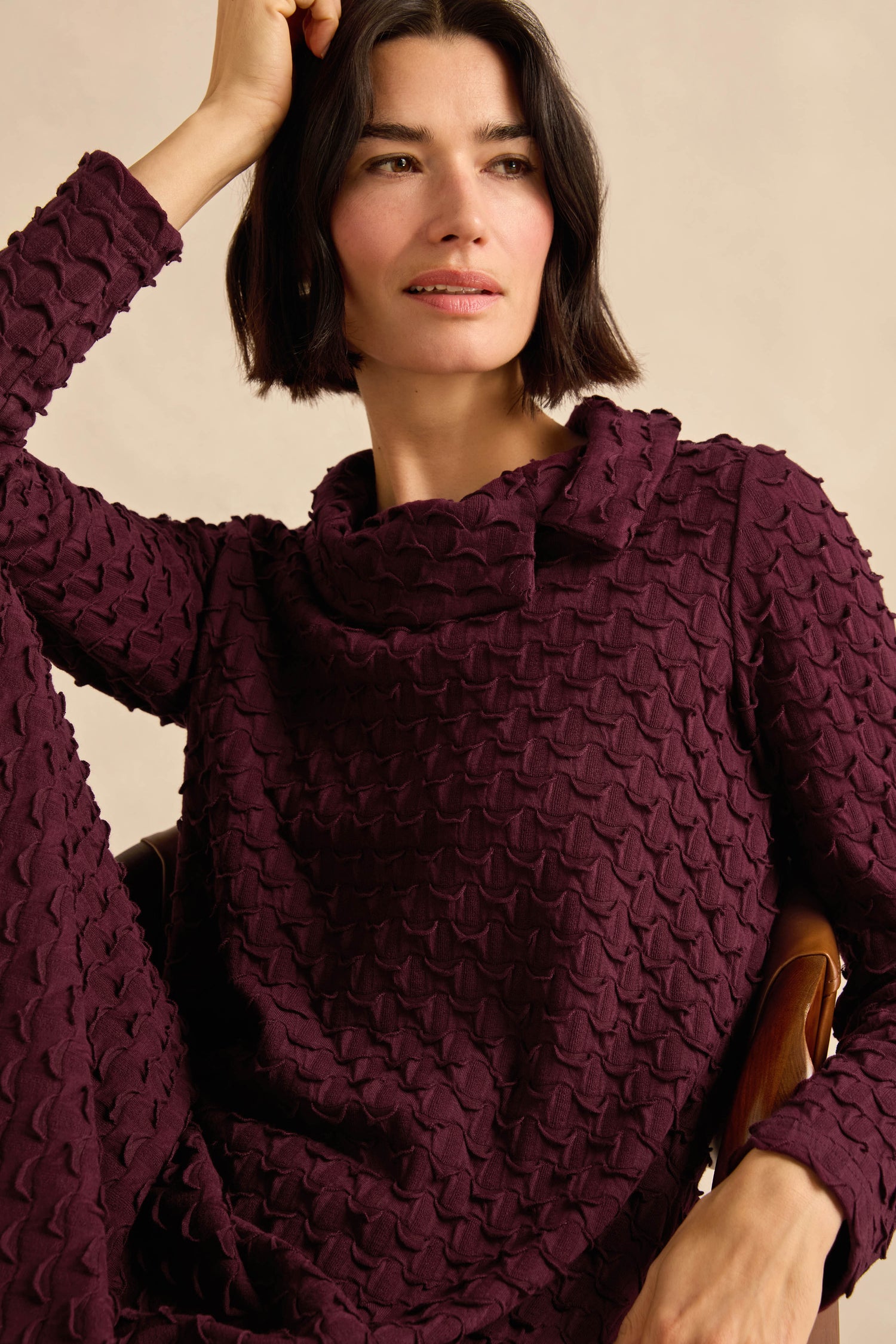 A woman with short dark hair wears the Origami Pocket Jersey Cowl Neck Dress in maroon, featuring a textured finish and wide collar, as she sits with her arm resting on her head against a neutral background.
