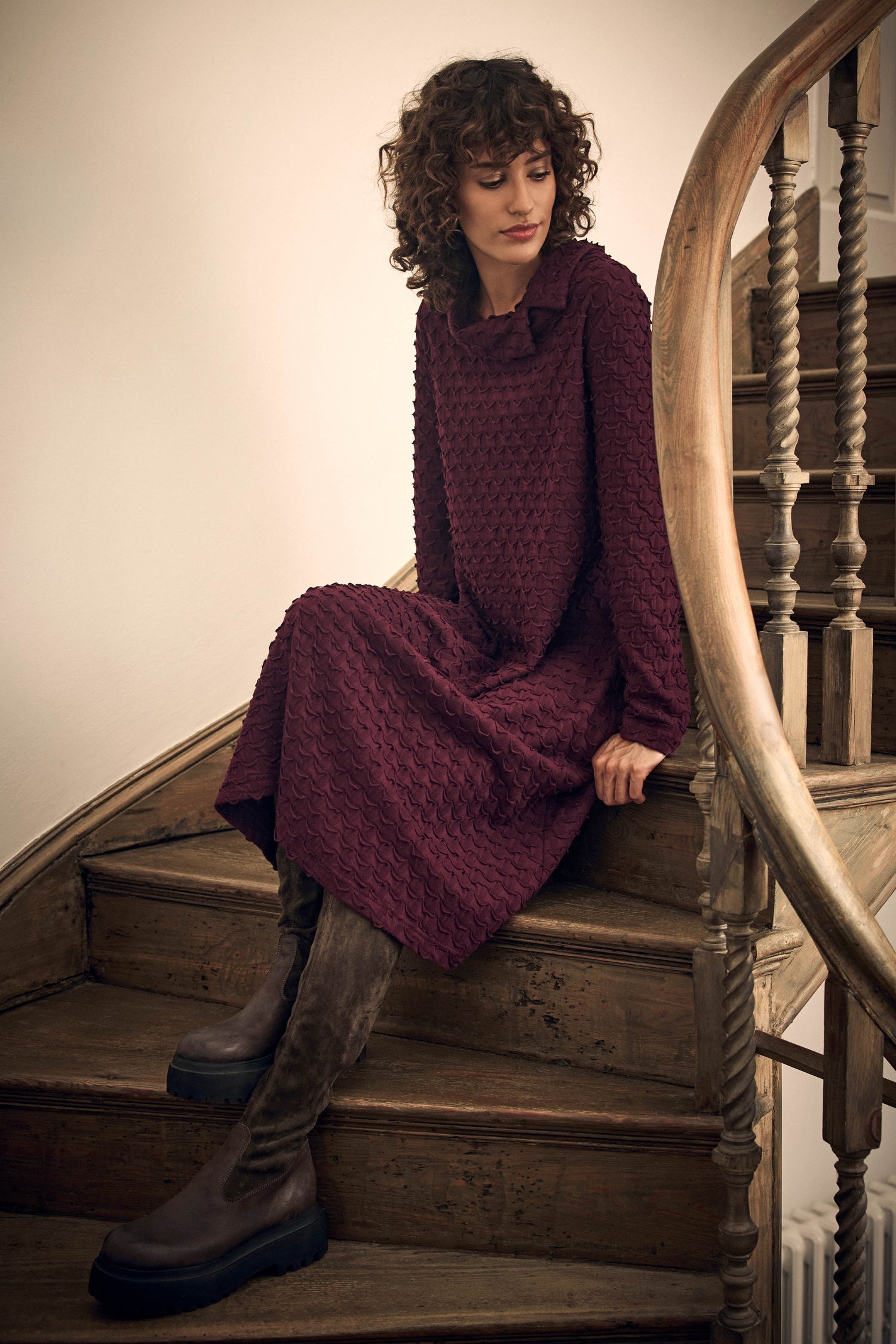 A woman in a dark purple, textured Origami Pocket Jersey Cowl Neck Dress sitting on wooden stairs, wearing tall dark boots.