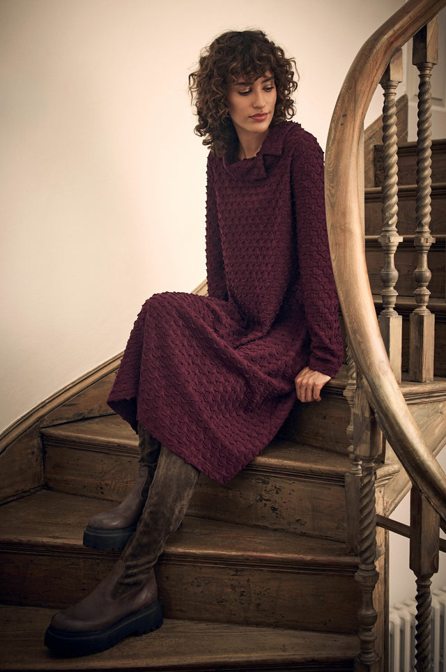 A woman in a dark purple, textured Origami Pocket Jersey Cowl Neck Dress sitting on wooden stairs, wearing tall dark boots.