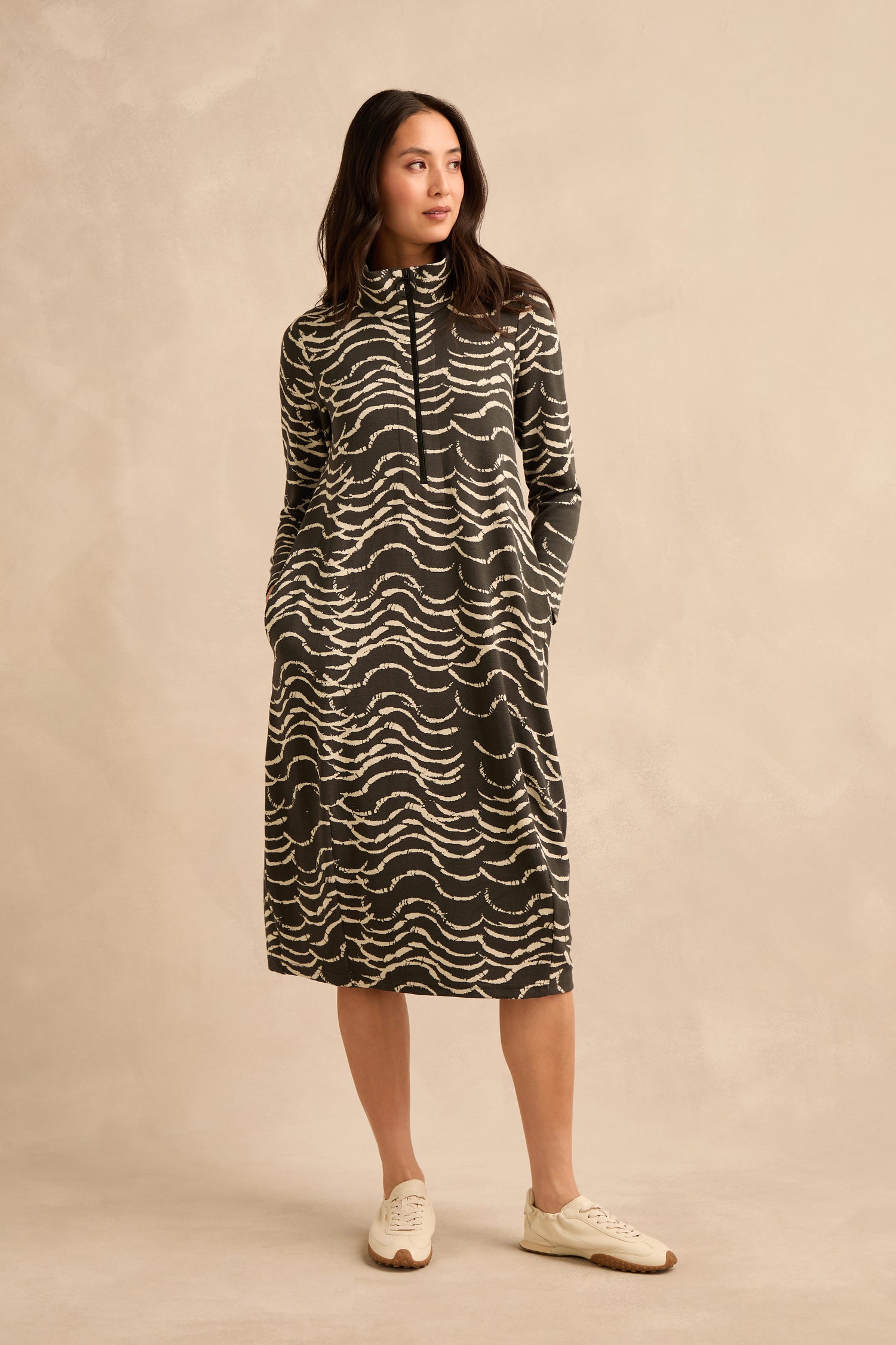 A person stands against a beige background wearing the Dancing Waves Jersey Zip Neck Dress—a long-sleeved, knee-length dress featuring a wavy black and white pattern—with white sneakers, hands in pockets, looking slightly to the side.