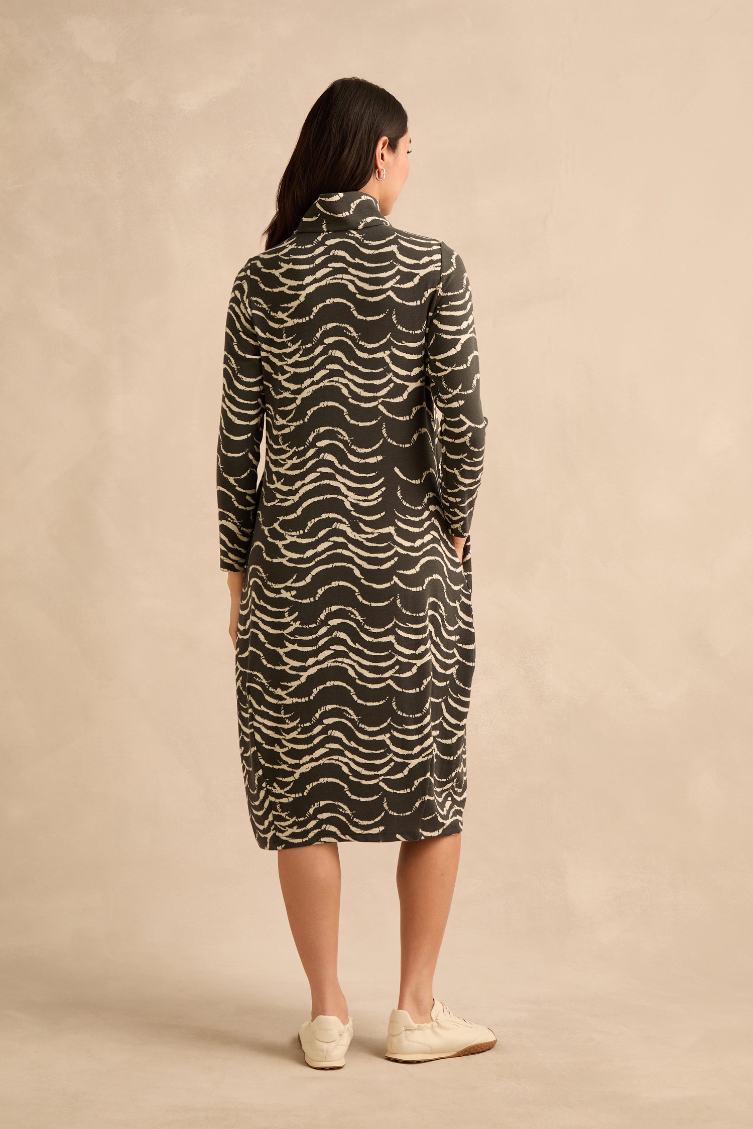 A woman stands facing away, wearing the Dancing Waves Jersey Zip Neck Dress featuring a knee-length, long-sleeve design with a wavy black and white pattern and a bubble silhouette that creates gentle volume, paired with cream-colored shoes against a neutral background.