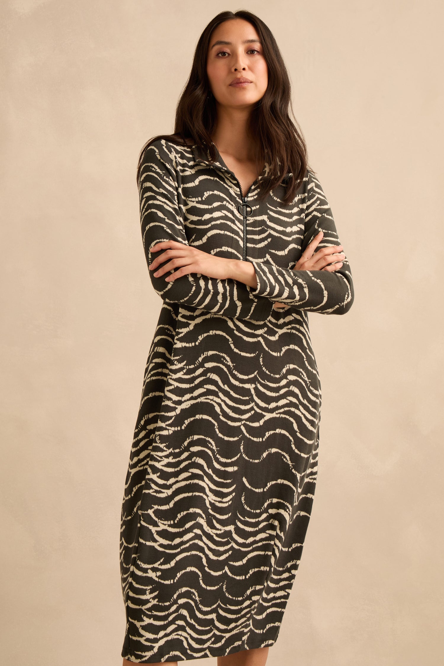 A woman stands with arms crossed, wearing the Dancing Waves Jersey Zip Neck Dress—a long-sleeved black midi dress featuring a white abstract wavy pattern and a refined bubble silhouette in stretch jersey—set against a plain beige background.