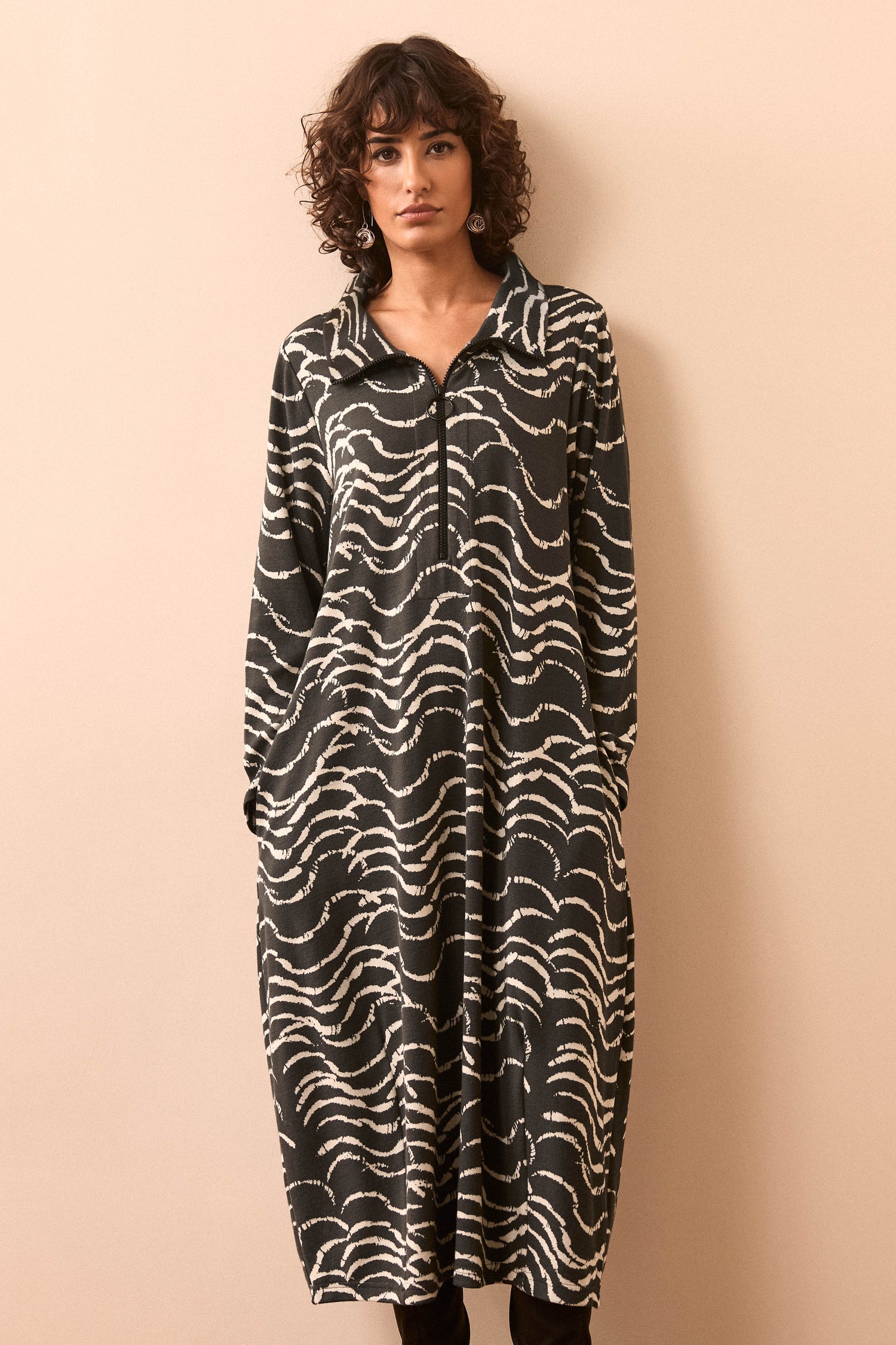 A woman models the Etched Wave Jersey Zip Neck Dress, a long-sleeved, loose-fitting piece featuring a black and white wavy stripe pattern and a subtle bubble silhouette, standing against a plain beige background.
