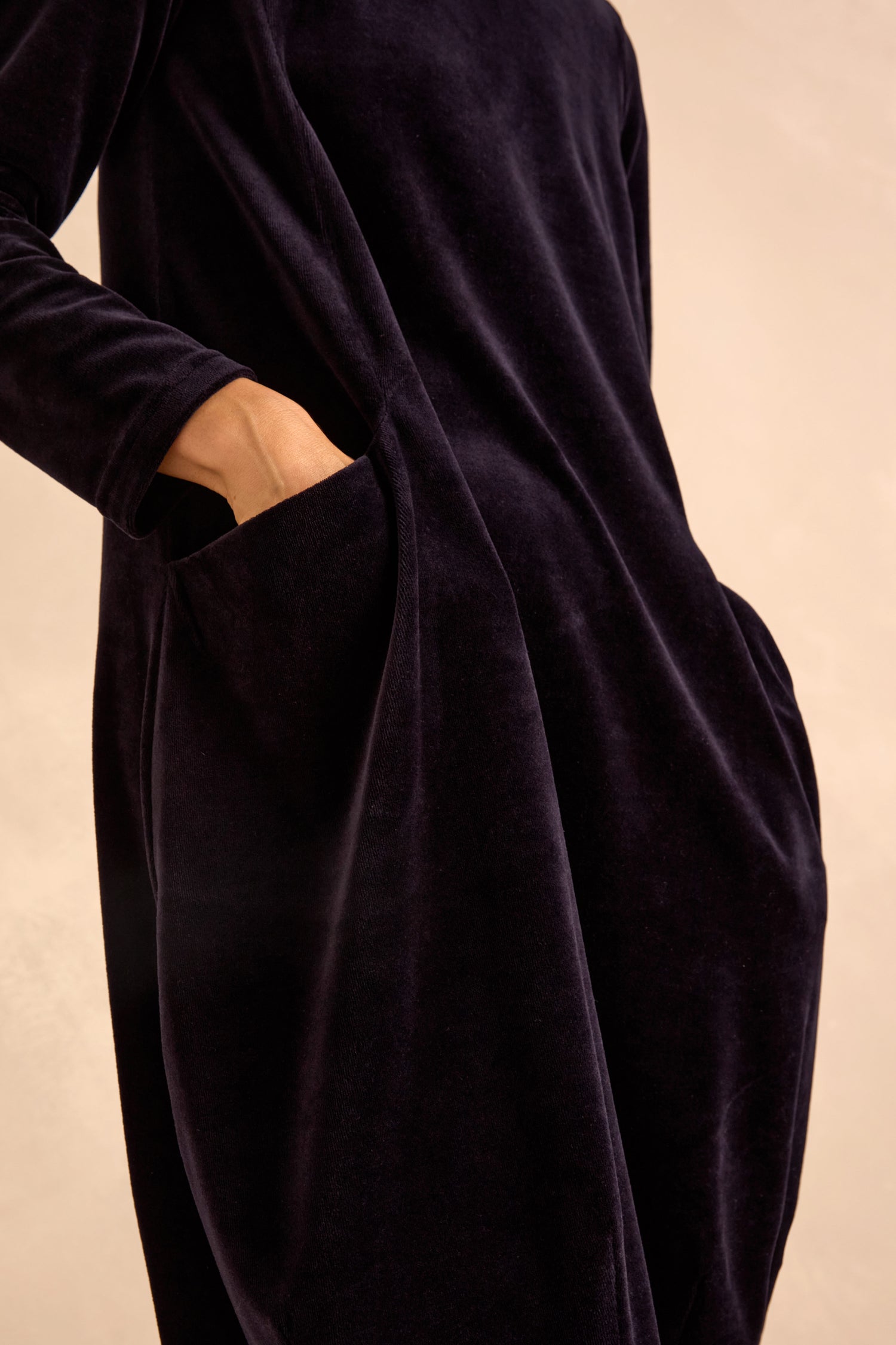 Model wears the Velvet Jersey Pocket Dress, a long-sleeved dark velvet style with discreet side pockets and a soft bubble silhouette, shown from shoulder to mid-thigh.