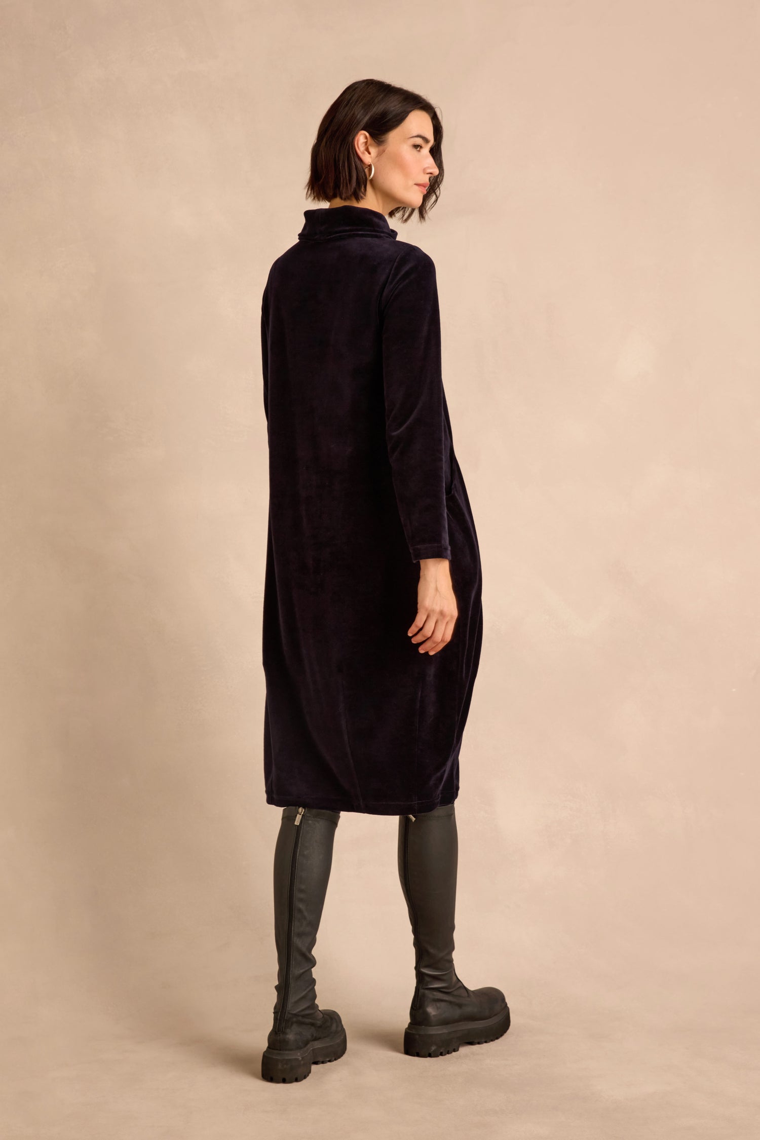 A woman stands sideways against a beige backdrop, wearing the Velvet Jersey Pocket Dress—a long black dress in velvet jersey with a subtle bubble silhouette—paired with black knee-high boots as she looks over her shoulder.