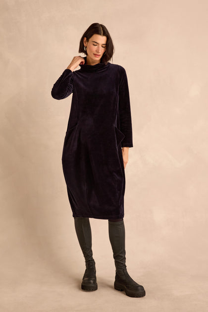 A woman wearing the Velvet Jersey Pocket Dress—a long-sleeve, dark velvet jersey dress featuring a cowl neck and side pockets—paired with dark leggings and black boots, stands against a neutral beige background.