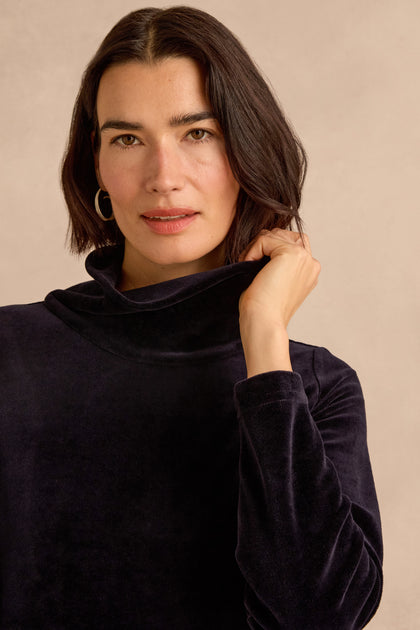 A woman with dark, shoulder-length hair stands against a plain beige background, wearing the Velvet Jersey Pocket Dress in navy with a subtle cowl neck and holding the collar with one hand.