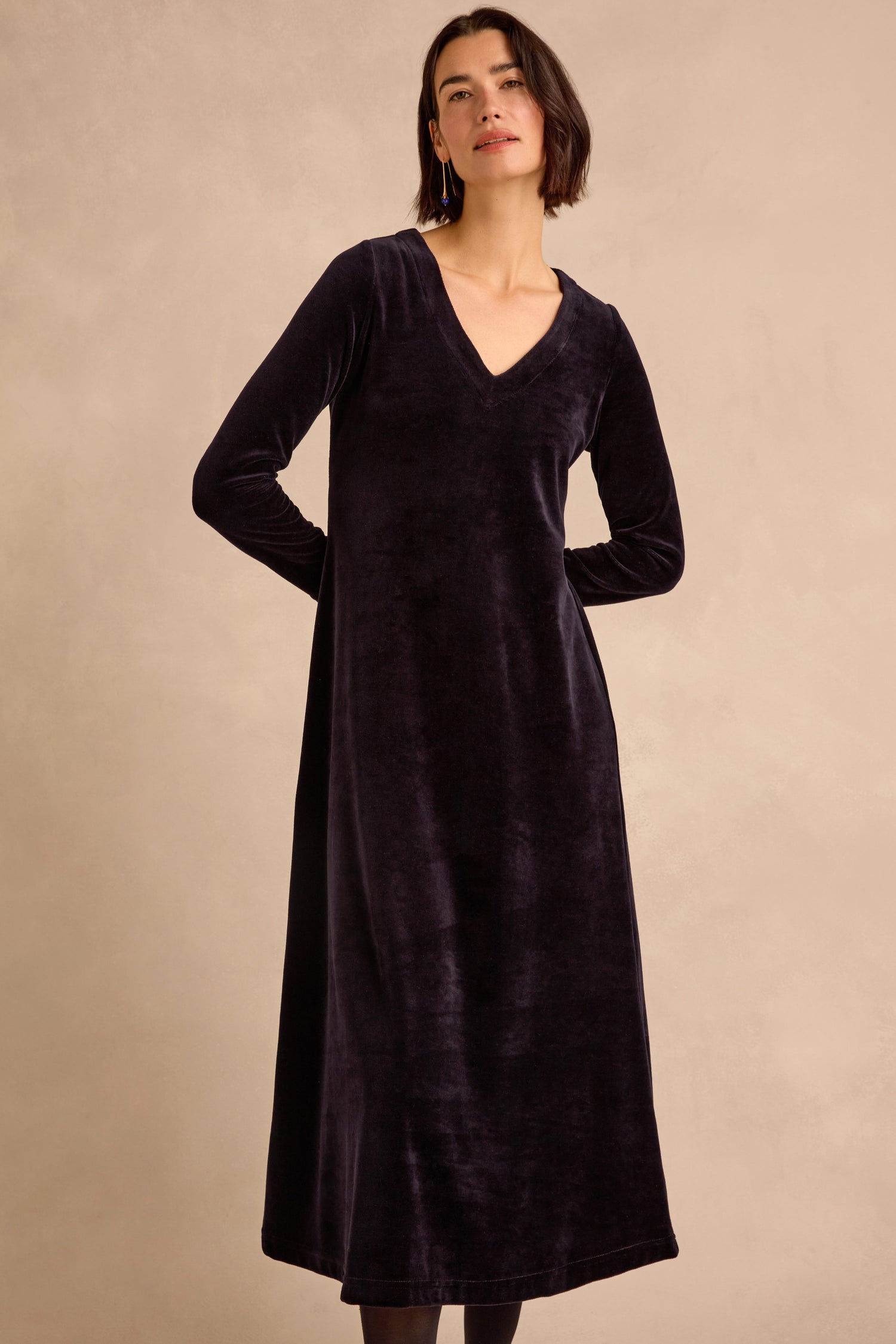 A woman stands against a plain beige background wearing the Night Velvet Jersey Maxi Dress—a long-sleeve, ankle-length black velvet jersey dress featuring a V-neckline and a flattering fit-and-flare silhouette.