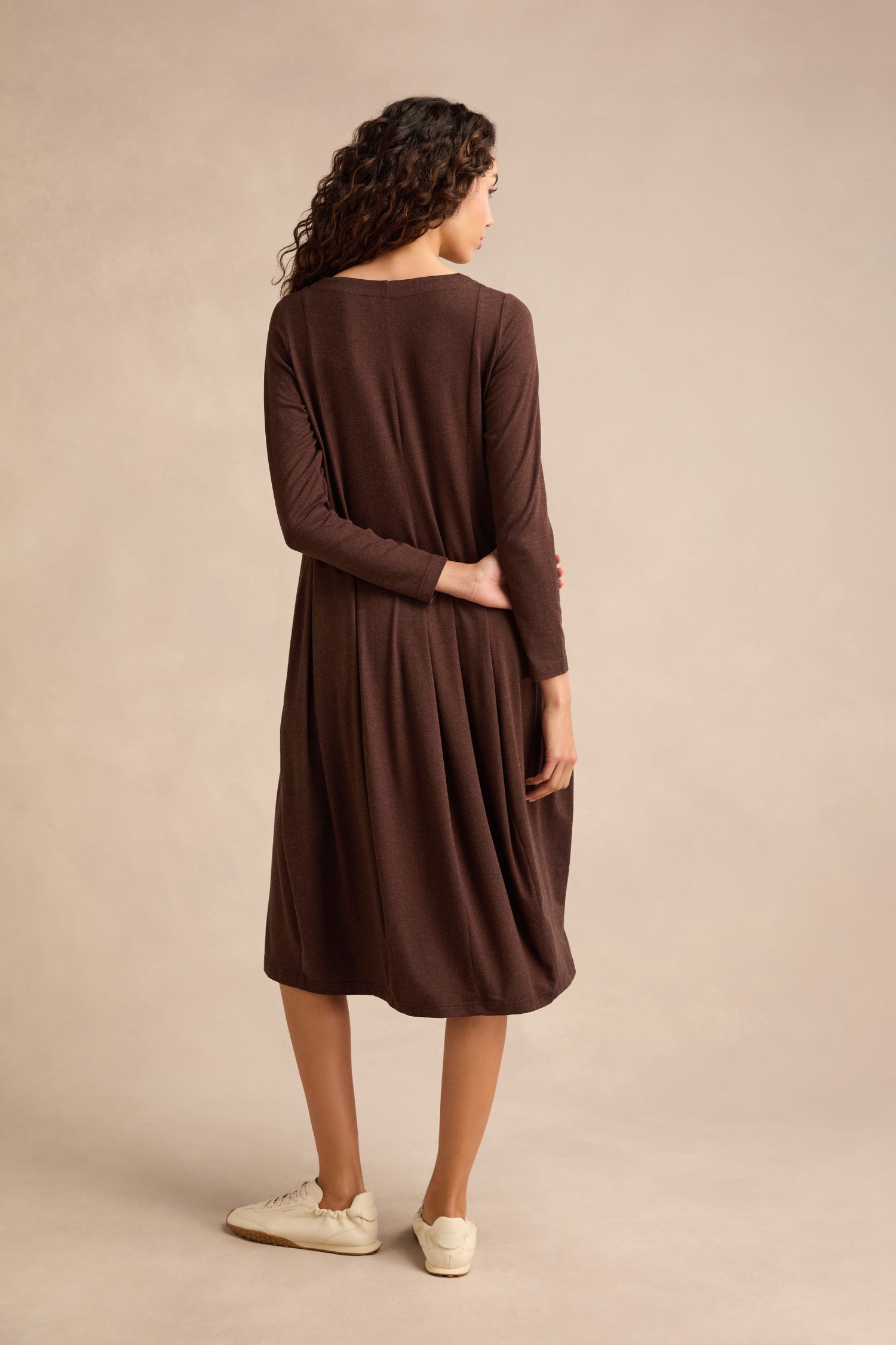 A woman with curly hair stands facing away, one hand behind her back, wearing the Soft Viscose Jersey Bubble Dress—a long-sleeve, knee-length dress in brown—paired with white sneakers.