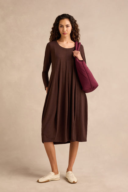 A woman stands against a plain background, wearing the Soft Viscose Jersey Bubble Dress in chocolate with long sleeves, paired with white sneakers and a maroon shoulder bag.