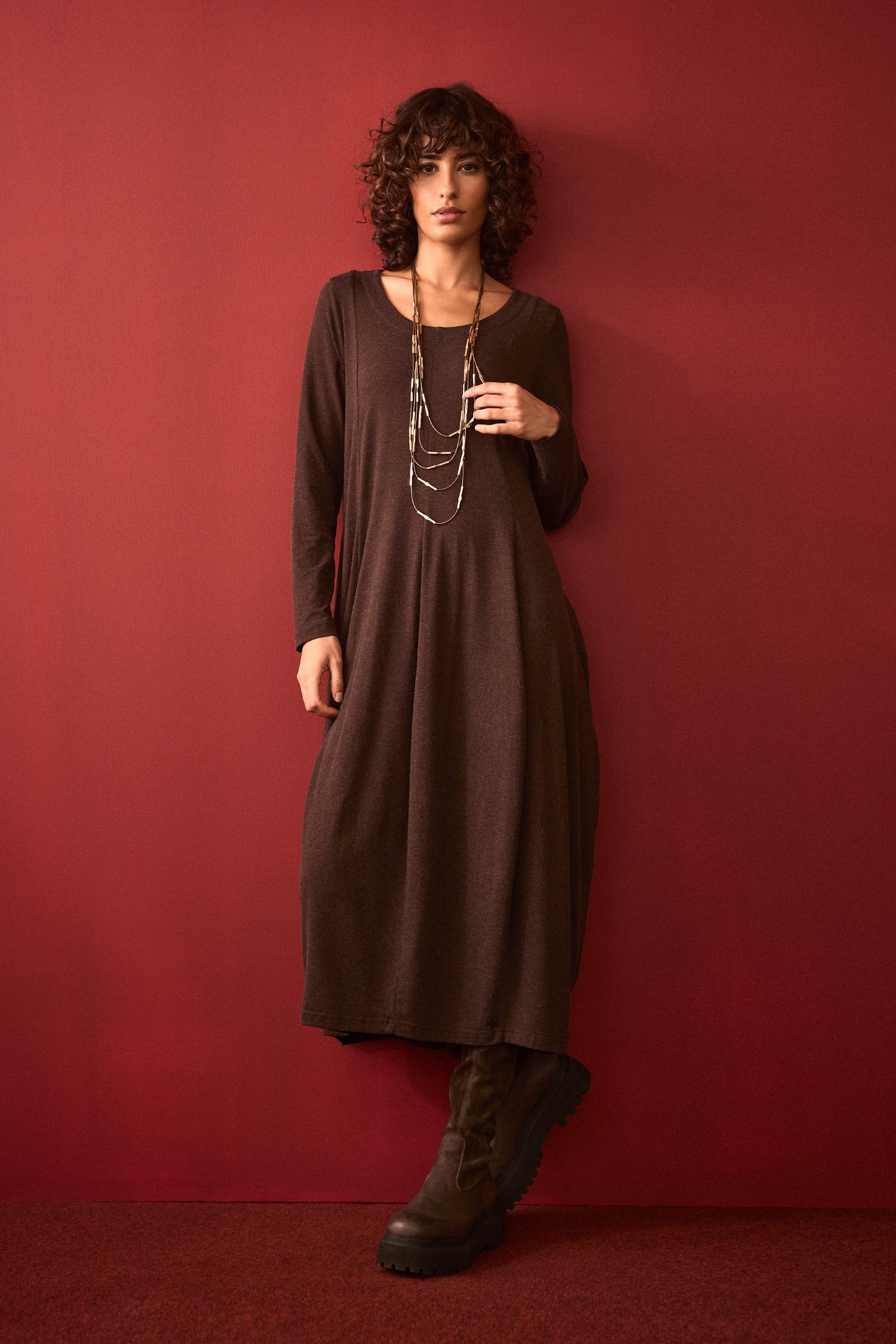 A woman with curly hair stands against a red wall, wearing the Soft Viscose Jersey Bubble Dress in brown, styled with a layered necklace and black boots. She has one hand in her pocket.