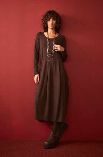 A woman with curly hair stands against a red wall, wearing the Soft Viscose Jersey Bubble Dress in brown, styled with a layered necklace and black boots. She has one hand in her pocket.