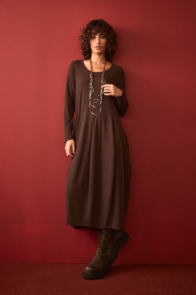 A woman with curly hair stands against a red wall, wearing the Soft Viscose Jersey Bubble Dress in brown, styled with a layered necklace and black boots. She has one hand in her pocket.