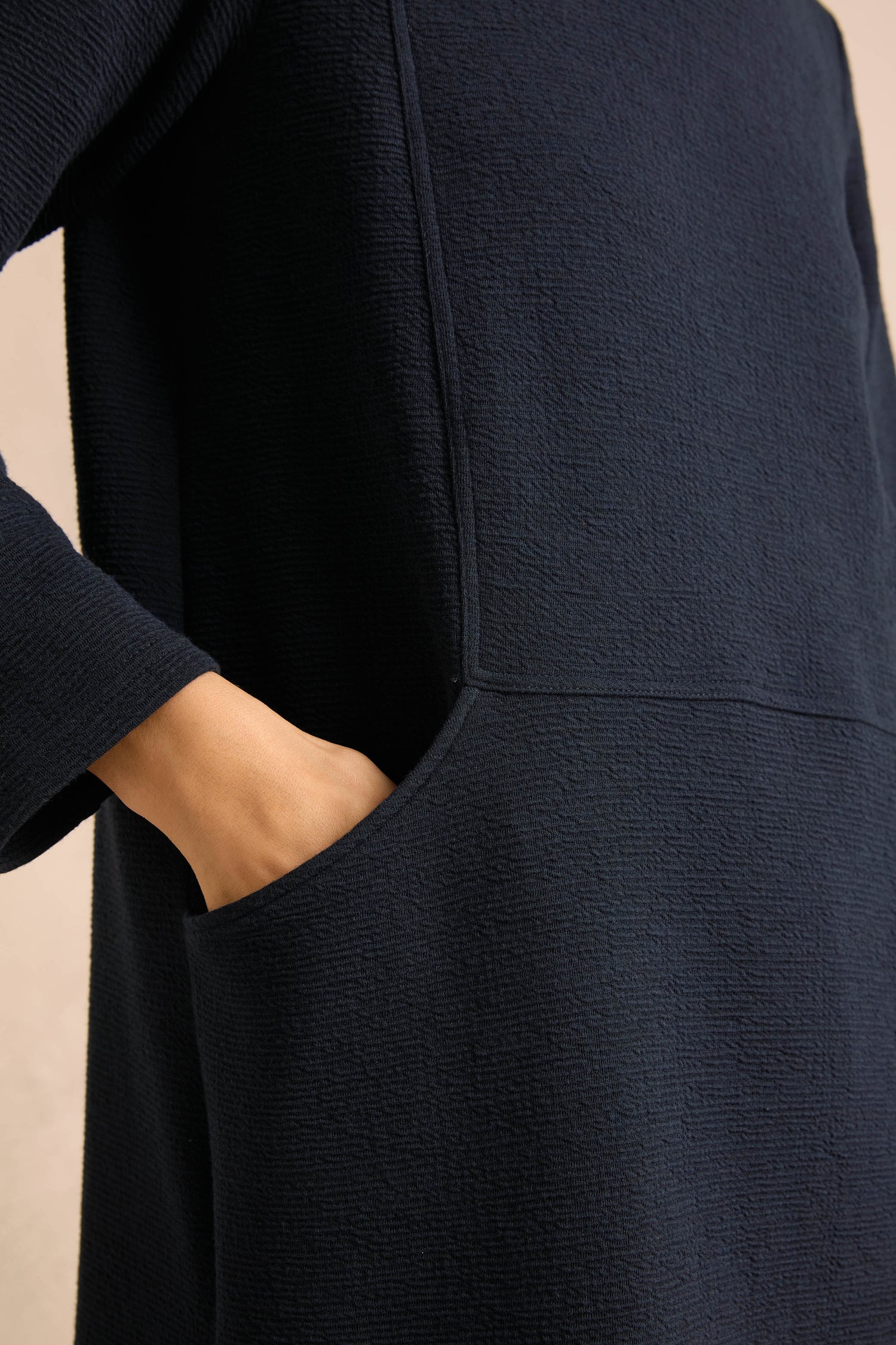 A person’s hand rests inside the front pocket of the textured, dark navy Double Jersey Crinkle Dress, highlighting its signature contrast cowl neck and unique details.