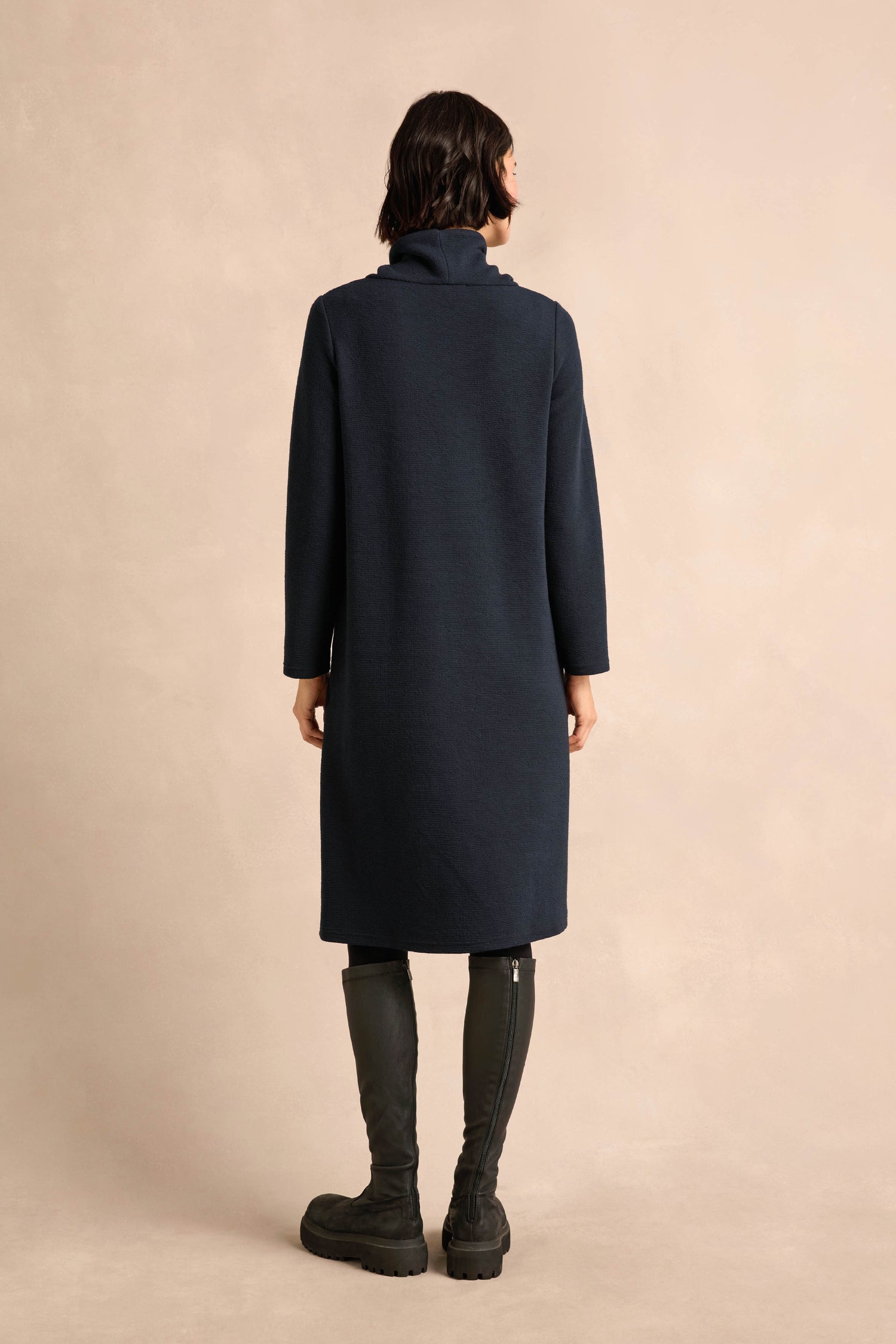 A person stands with their back to the camera, dressed in the Double Jersey Crinkle Dress in navy with long sleeves, paired with tall black boots, set against a plain beige background.