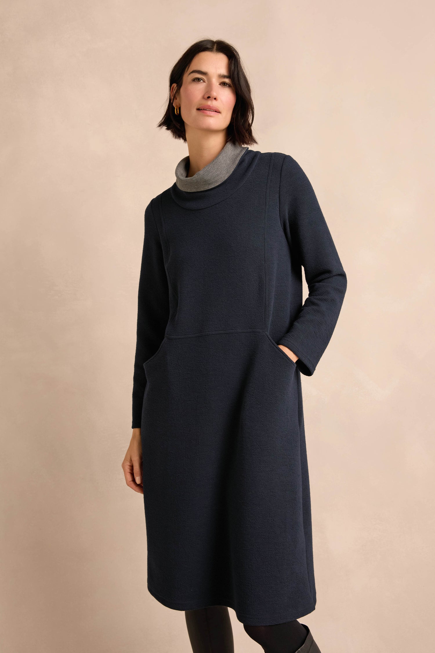 A woman stands against a plain beige background wearing the Double Jersey Crinkle Dress, a knee-length, long-sleeve design featuring a gray contrast cowl neckline.