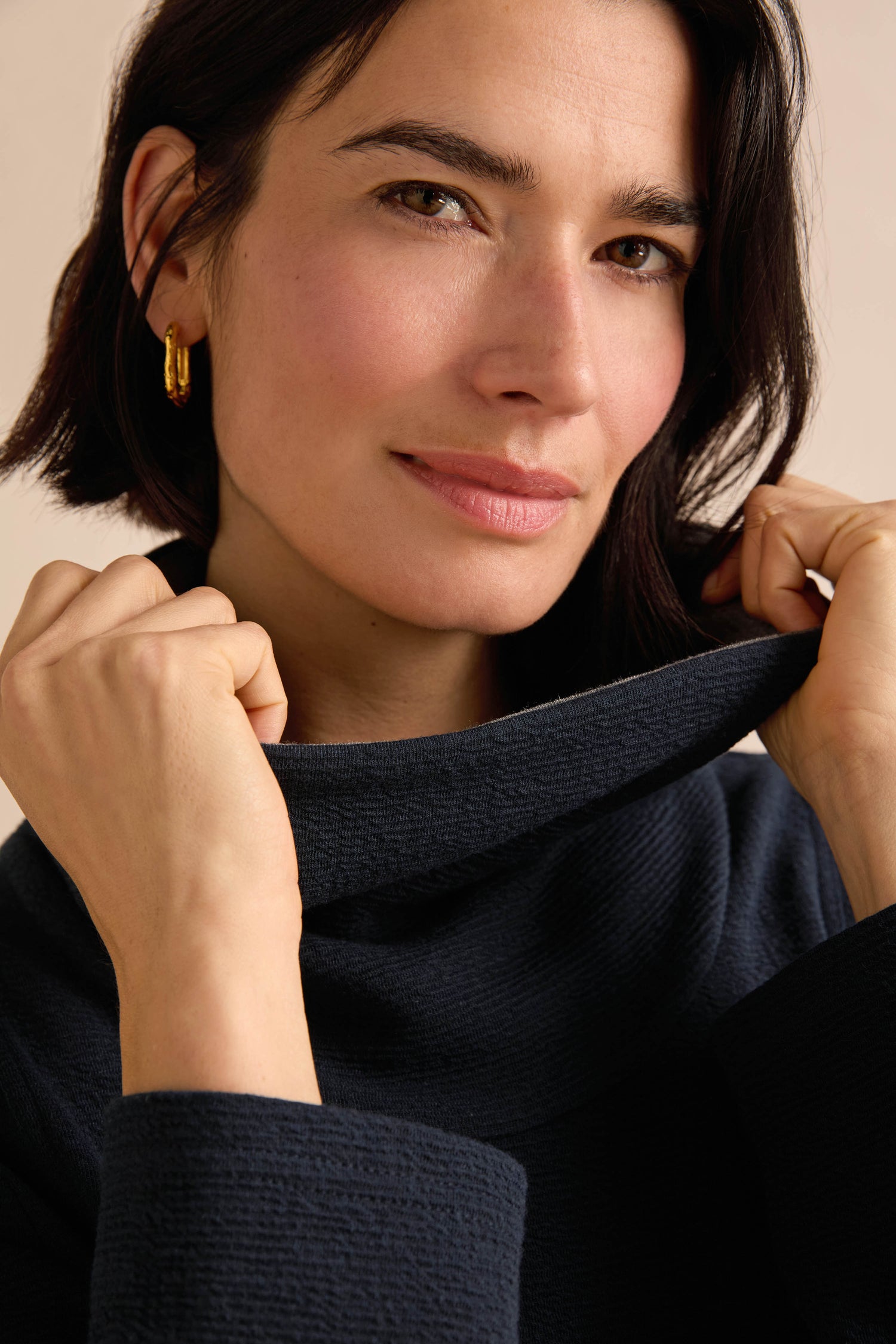 A woman with short dark hair wears the Double Jersey Crinkle Dress, featuring a navy blue textured fabric and a contrasting cowl neck. She holds the collar with both hands and looks at the camera with a slight smile.