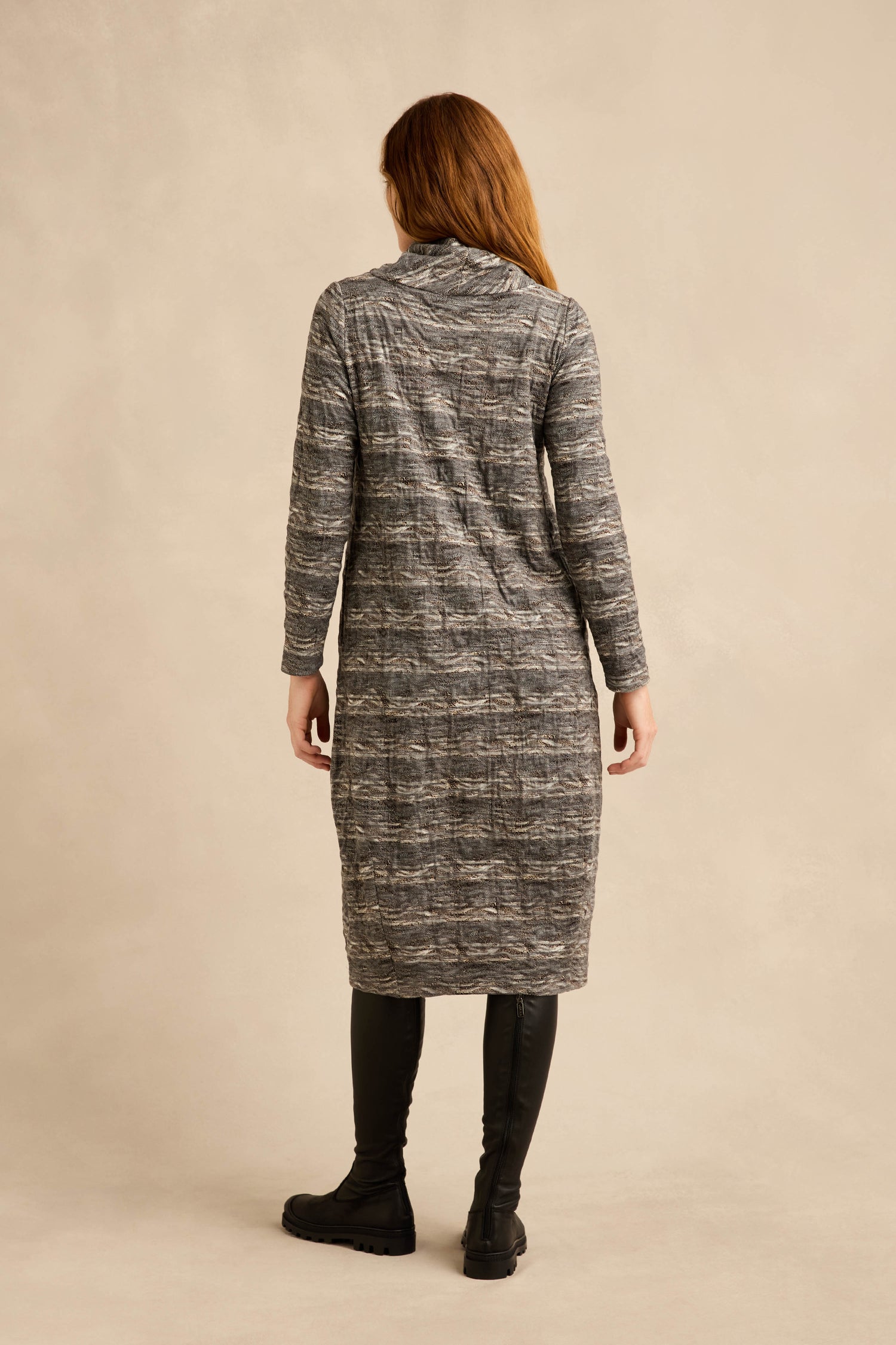 A person stands facing away, wearing a long-sleeved, knee-length gray Abstract Ripple Jersey Bubble Dress with a textured jersey finish and black leggings with black boots, against a beige background.