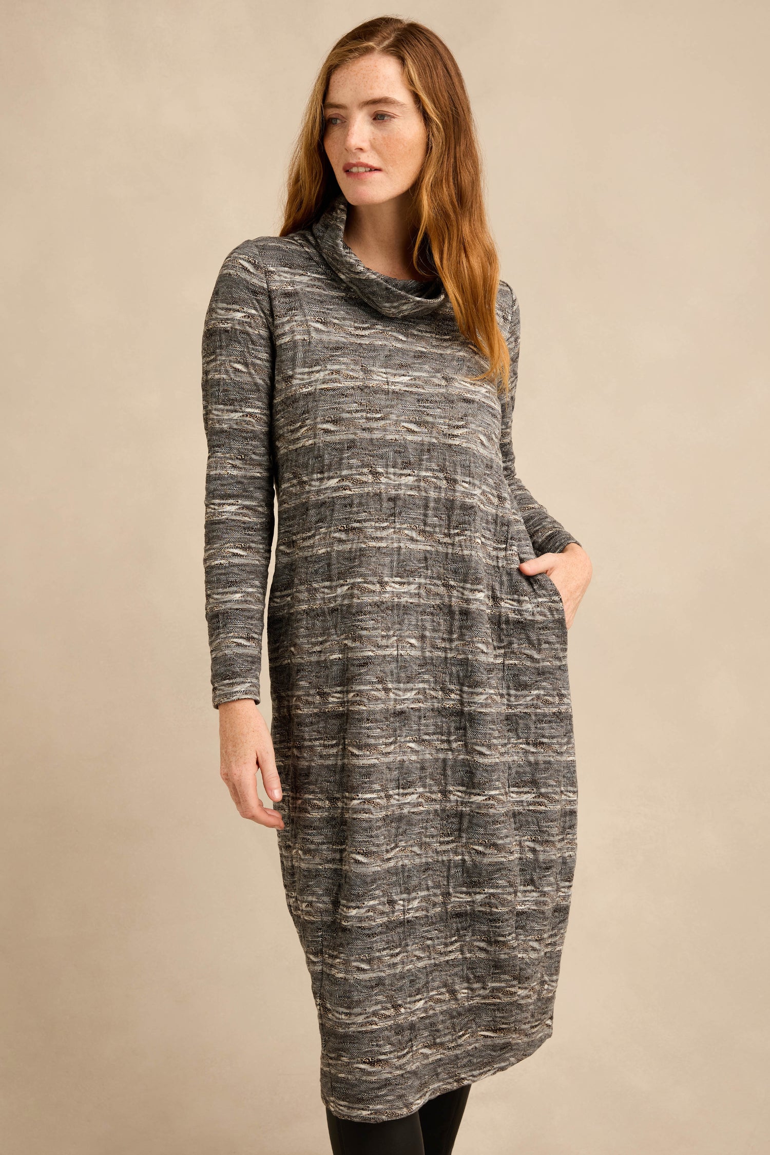 A woman stands against a plain beige background, wearing the Abstract Ripple Jersey Bubble Dress—a long-sleeved, gray patterned dress with a cowl neck, side pockets, and a subtle bubble silhouette.