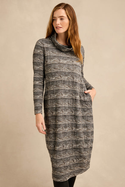 A woman stands against a plain beige background, wearing the Abstract Ripple Jersey Bubble Dress—a long-sleeved, gray patterned dress with a cowl neck, side pockets, and a subtle bubble silhouette.