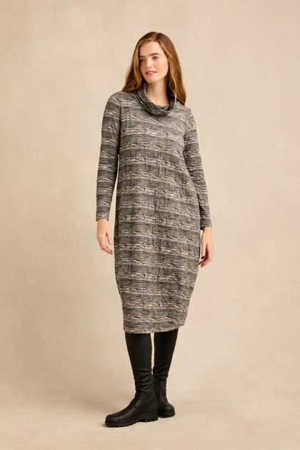 A woman stands against a plain beige background wearing the Abstract Ripple Jersey Bubble Dress in gray, featuring long sleeves, a subtle bubble silhouette, and paired with black knee-high boots.