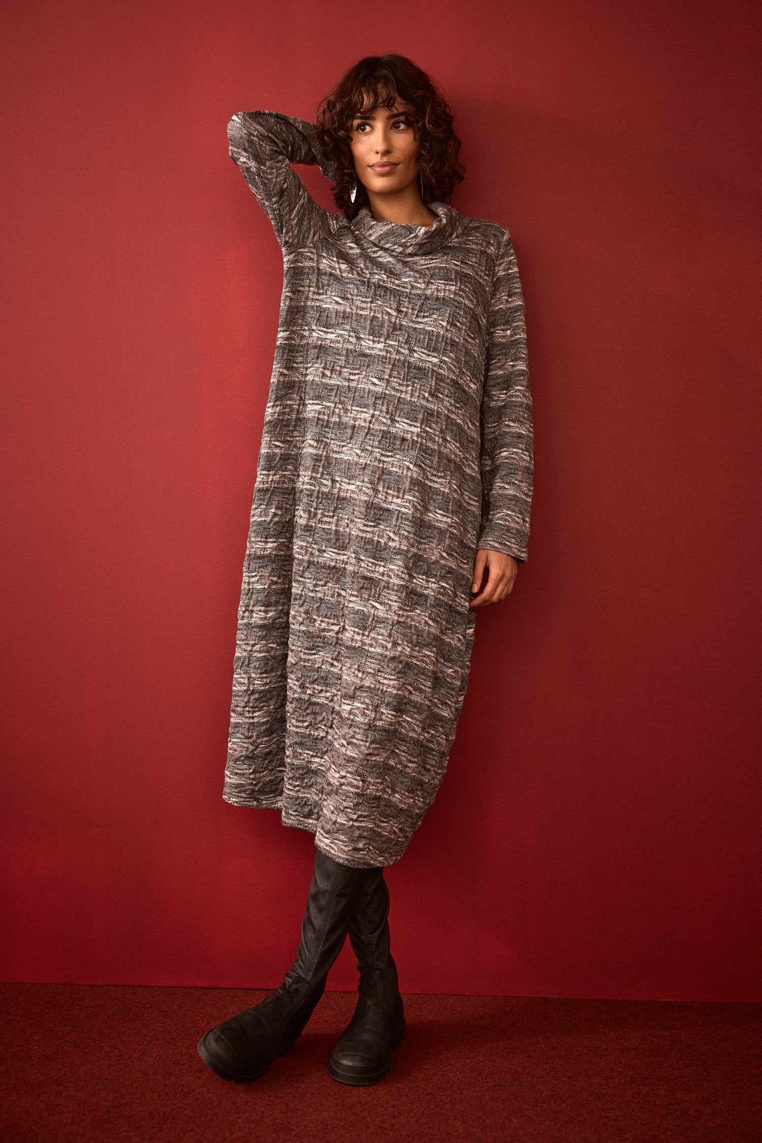 A person stands against a red wall with one hand behind their head, wearing black boots and the Abstract Ripple Jersey Bubble Dress. The textured jersey fabric and bubble silhouette add depth and dimension to this striking look.