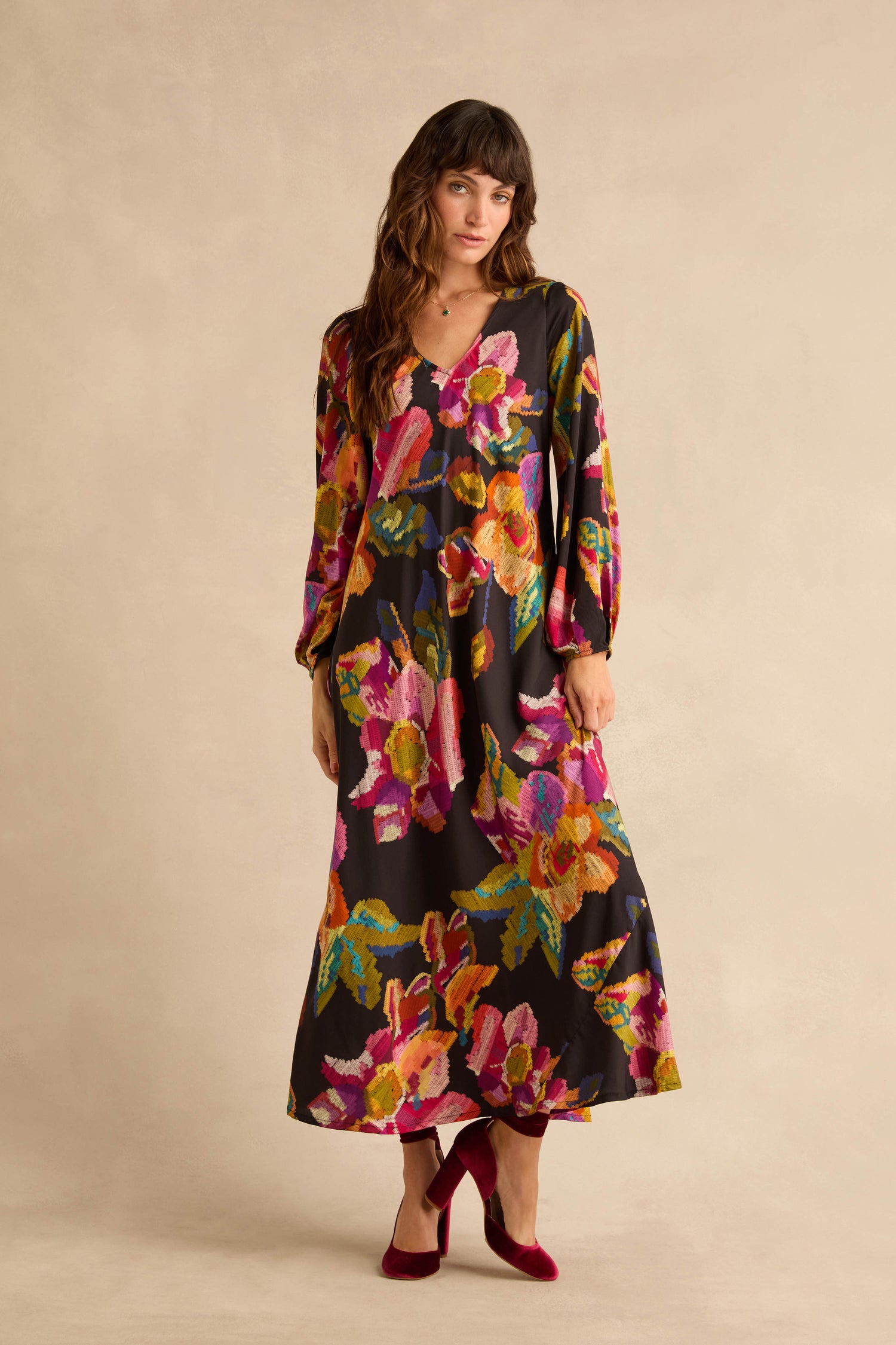 A woman wearing a Pixelated Floral Flared Dress in pure viscose with a black background and colorful patterns, paired with burgundy velvet block-heel pumps.