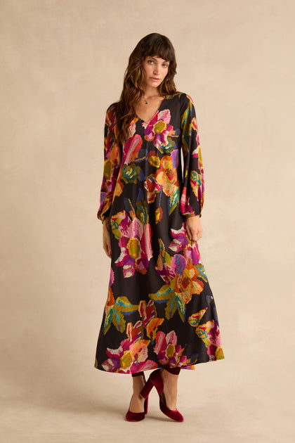 A woman wearing a Pixelated Floral Flared Dress in pure viscose with a black background and colorful patterns, paired with burgundy velvet block-heel pumps.
