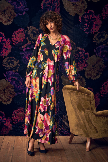 A model stands next to an armchair wearing a Pixelated Floral Flared Dress, a floor-length, flared maxi dress featuring a colorful, pixelated floral pattern.