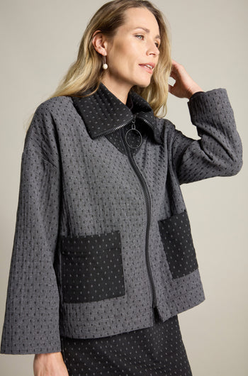 A woman with long blonde hair is wearing a Square Jacquard Boxy Jacket. This contemporary style, grey patterned zip-up jacket features large black pockets. The textured boxy fit jacket complements her raised hand as she gazes slightly to the side.