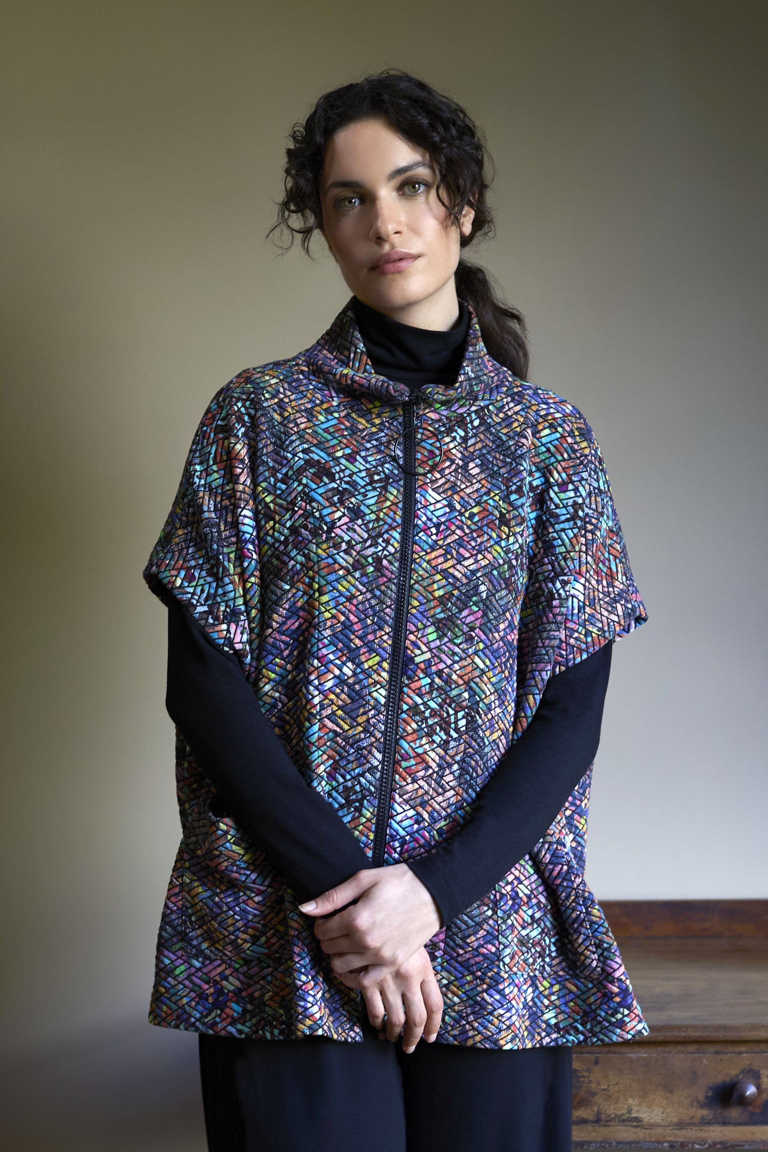 A woman with curly dark hair wears a vibrant, multi-coloured Diamond Jacquard Jersey Jacket over a black turtleneck, standing with hands clasped in front of a plain background.