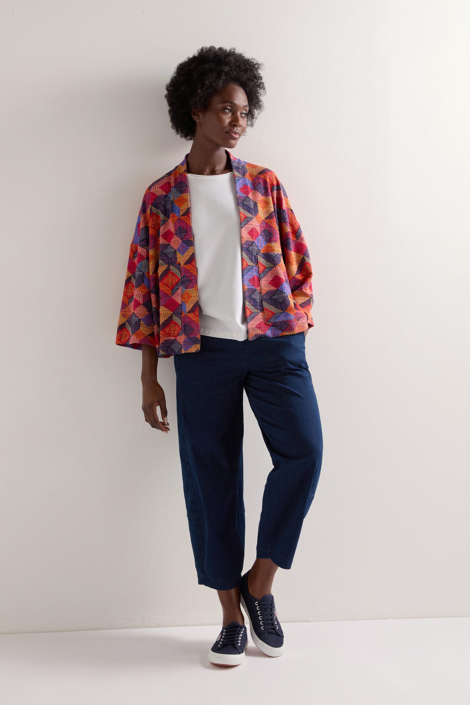 Wearing the Bold Cubist Print Jacket over a white shirt, paired with blue pants and black sneakers against a white wall, creates an ensemble showcasing contemporary style with colorful geometric patterns.