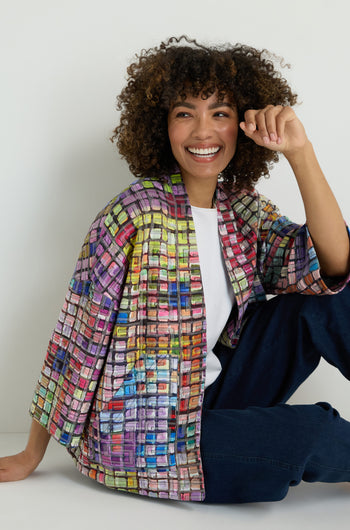 A woman wears the Coloured Glass Tiles Jersey Jacket—a lightweight blue top and pants set with vibrant glass tile patterns—paired with white sneakers against a plain light background.