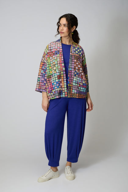 A woman wears the Coloured Glass Tiles Jersey Jacket—a lightweight blue top and pants set with vibrant glass tile patterns—paired with white sneakers against a plain light background.