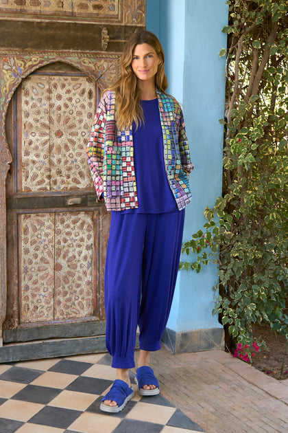 A woman stands by greenery, smiling in the Coloured Glass Tiles Jersey Jacket over a royal blue top with matching loose pants and blue sandals. The vibrant, lightweight jacket adds a lively touch to her look.